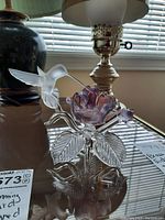 Image showing a frosted glass hummingbird hovering against a purple flower with clear textured leaves on a reflective glass surface.