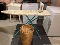 Ironing board with colorful floral padded cover, teal metal legs, electric iron resting on board, laundry appliances and sink in background.