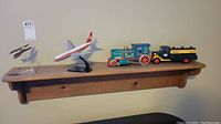 Four model items on wooden shelf: two model airplanes, Hess scale truck, tin train