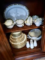 Photo of Noritake Warrington pattern teacups with saucers, stacked plates and salt and pepper shakers inside a wooden cabinet.