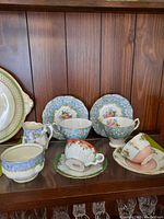 View of multiple teacups and saucers displayed on a wooden shelf with a wood panel backdrop. Includes multiple floral designed teacups and saucers, some matching, some unique.