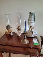 Front view of three oil lamps on wooden table showing pottery base lamp, pressed glass base lamp, brass base lamp, and painted brass candle holder with hurricane.