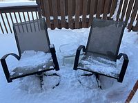 Two outdoor swivel bottom chairs with black metal frames and mesh seats, and a small round glass top side table covered in snow on a snowy deck.