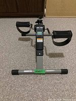 Front view of pedal exerciser showing foot pedals with straps and digital monitor screen with warning label.