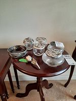 Photo showing five clear glass dresser jars arranged on wooden table, each topped with decorative pewter lids with floral and leaf designs. Also visible are a pewter rose and a small pewter tray with grapevine motif.