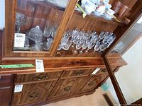 Wooden cabinet with shelves displaying multiple crystal glasses and dishes including whisky glasses, water goblets, covered butter dish, candle holders, and shot glasses