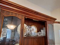 Wood cabinet with crystal glassware including a clear pinwheel crystal pitcher, decanter sugar bowl, swan bell, and various plates visible inside shelving.
