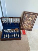 Wooden case open on floor showing silverware inside including spoons, forks and serving pieces, with floral box and red-white box beside it