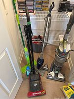 Photo showing five cleaning items: Hoover Air Pro vacuum, Dirt Devil Roommate vacuum, Shark steam mop, green Libman floor mop and mop bucket, plus mop cleaning solution bottle.