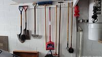Photo showing hanging yard tools: shovels, brooms, dustpan, hand tiller, spade, and edger on concrete wall.