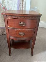 Front view of wooden bedside table showing two drawers with metal oval handles and lower shelf. Carved detailing beneath the top drawer and along legs visible. Table finish is reddish-brown wood with surface wear.