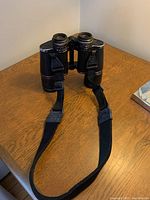 View of entire binoculars with attached black strap on wooden surface.