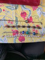 Four bracelets displayed on a floral patterned cloth, showing varied materials and styles including amber stones, simulated pearls, and colorful beads.