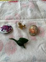 Four brooches: a purple and pink painted oval brooch, a gold-colored goose with green eye brooch, an oval enamel brooch with woman painting, and a green jade goose brooch on floral fabric background.