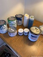 Photo showing nine items including five paint cans or jars, two small sample jars, and two squeeze bottles on a wooden table under warm light.