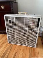 Front view of the white electric standing fan with a protective grille and power cord on a wooden floor.