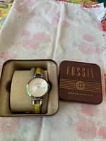 Photo of the Fossil watch inside its original beige cushion and the brown metal case with 'FOSSIL WORLD AIRWAYS' branding visible.