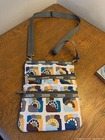 Full view of the LeSportsac cross body bag on wooden table showing adjustable gray strap and three zippered pockets with multicolor geometric fabric.