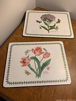 Two rectangular botanical luncheon placemats with flower illustrations and green ivy border, one featuring tulips, the other with pink phlox, butterflies present