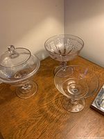 Three pressed glass comports displayed on a wooden surface, one with lid, one patterned, and one clear no pattern.
