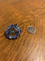 Close-up photo of circular Sherman brooch with blue and clear rhinestones lying on wooden table, quarter coin next to brooch for size reference