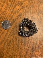 Signed Sherman brooch shown next to a coin for size reference. Black and smoky rhinestones in a triangular wreath design on wooden surface.