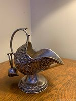View of silver-colored decorative vessel showing floral engravings, curved spout, arched handle with scoop hanging