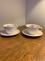 Two white ceramic Spode Jewel teacups each on a matching saucer displayed on wooden surface. Both show embossed details and pink rose floral decoration inside the cups.
