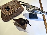 Photo of the small dark wooden bird carving, the larger painted bird carving on base, and the vintage fishing creel basket showing overall lot items