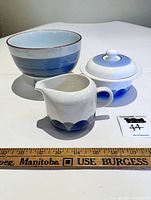 Three ceramic pieces displayed including a sugar bowl with lid, a creamer pitcher both with blue Artica pattern, alongside a blue Japanese bowl with a white interior. A ruler shows measurement for scale.
