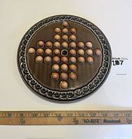Top view of round ceramic Solitaire game board with wooden pieces arranged in a cross pattern, ruler for scale shows approx 15 inches diameter.