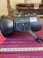 Side view of the Courtenay SolaFlash 1000S black metal lighting flash head with attached handle and mounting bracket, placed on a red cloth with a measuring ruler beneath.