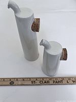 Two white ceramic oil and vinegar bottles with cork stoppers and angled spouts next to ruler for size reference.