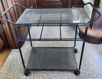 Full view of vintage black metal serving trolley with two shelves and wheels on carpeted floor.