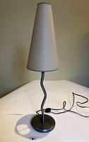Front view of tall lamp with conical white paper shade and squiggly black stem, placed on white surface against beige wall.