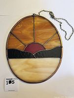 Front view of the oval stained glass pendant showing the sunset design with warm amber rays, black hills, maroon sun, and cream base along with the hanging chain.