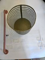Top-down photo of a brass-colored perforated metal wastebasket, pink plastic towel bar, and two clear lucite towel bars arranged on a white surface.