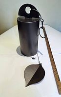 Front view of the black finished cylindrical steel bell with the hanging loop and attached leaf-shaped windcatcher on a white table.