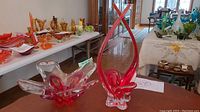 Lot on display showing the red and clear chalet style hand-blown art glass vase on a wood table with additional art glass pieces in the background.