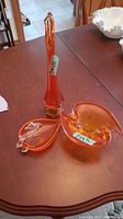 Three orange art glass pieces displayed on a wooden table, including a tall vase and two dishes