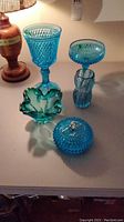 Blue glass compote, diamond pattern goblet, leaf-shaped bowl, and small vase on white table with lamp in background