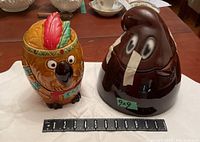 Front view of both ceramic cookie jars with measuring tape for scale. Owl jar shows colorful feather headband and facial details, Nabisco jar is dark brown with cartoon eyes.