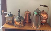 The full set of four copper tone kettles and the wood bodied coffee grinder arranged on a table