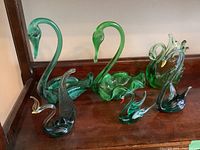 Six green glass swan figurines displayed on wooden shelf, all visible in varying sizes and green tones.