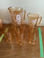 Front view of eight amber glassware pieces including two jugs with white floral motifs and six matching amber glasses.