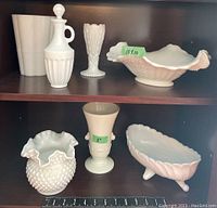 Full view of seven assorted milk glass pieces arranged on wood shelf including vases, bowls, and cruet.