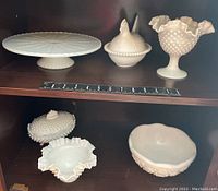 Six pieces of milk glass displayed on wooden shelves with measuring rulers for scale. Includes a large cake tray, two lidded containers, a footed compote, two candy dishes, and a bowl in hobnail pattern and smooth finishes.