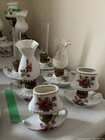 Six miniature ceramic lanterns with floral decals and gold trim, set on a white table. Varying styles of lantern tops including clear glass and ceramic shades.