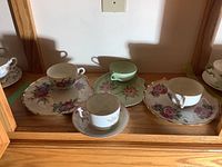 Three tea snack sets and one cup and saucer inside a wooden cabinet, showing floral patterns and gold accents on the cups and plates.