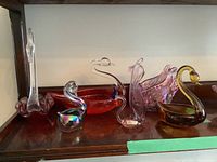 Six glass swan figurines arranged on a wooden shelf, various colors and styles visible including red, iridescent, blue-headed, pink, amber and clear glass swans.